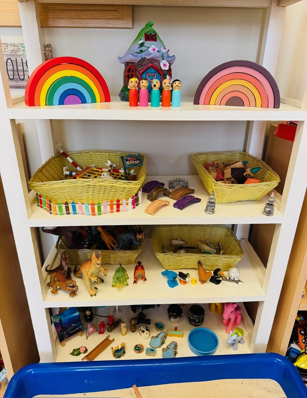 A calm, ordered shelf of rainbow wooden toys, peg-doll families and baskets of miniatures in the playroom