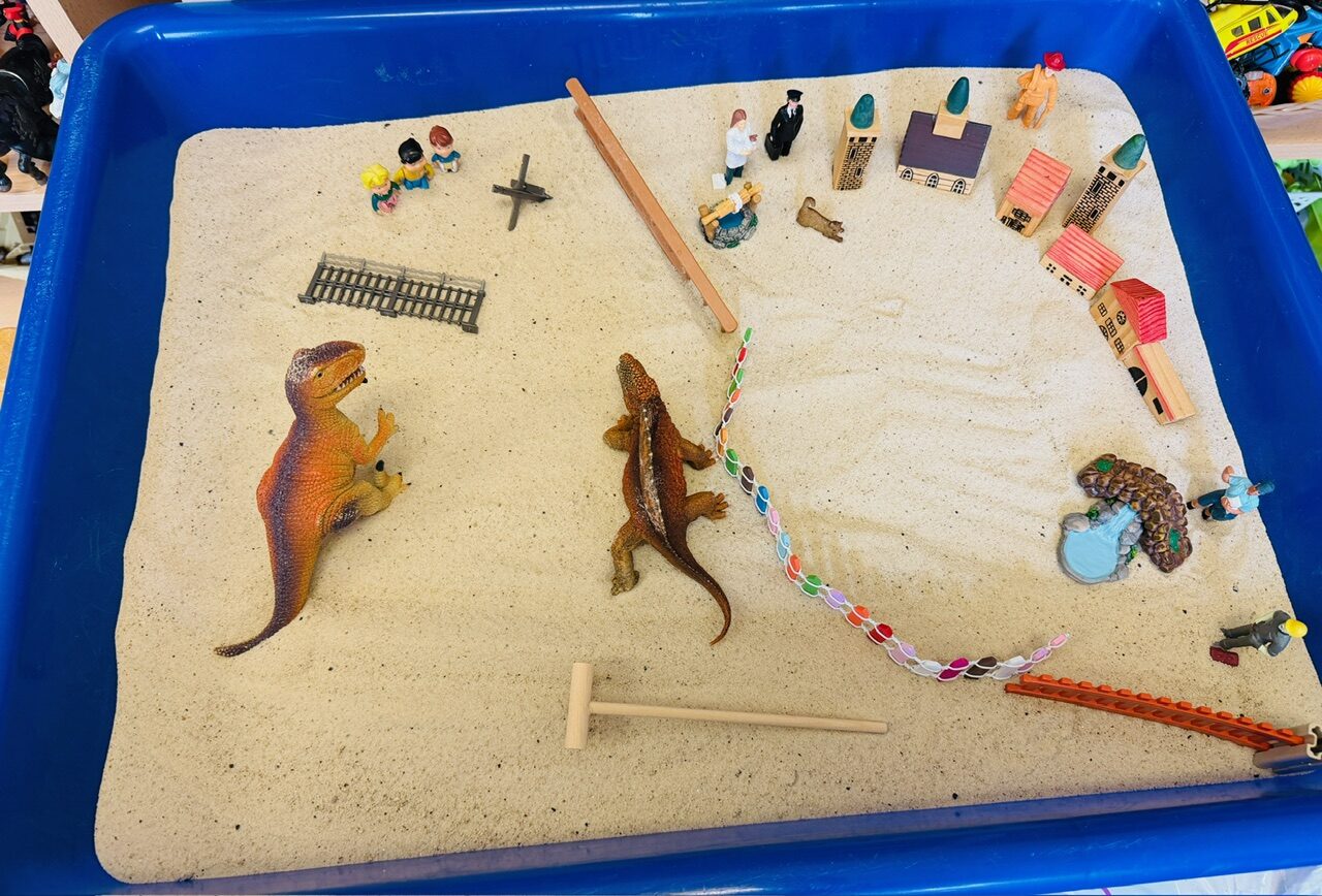 A blue sand tray arranged with dinosaurs, tiny houses and a winding path